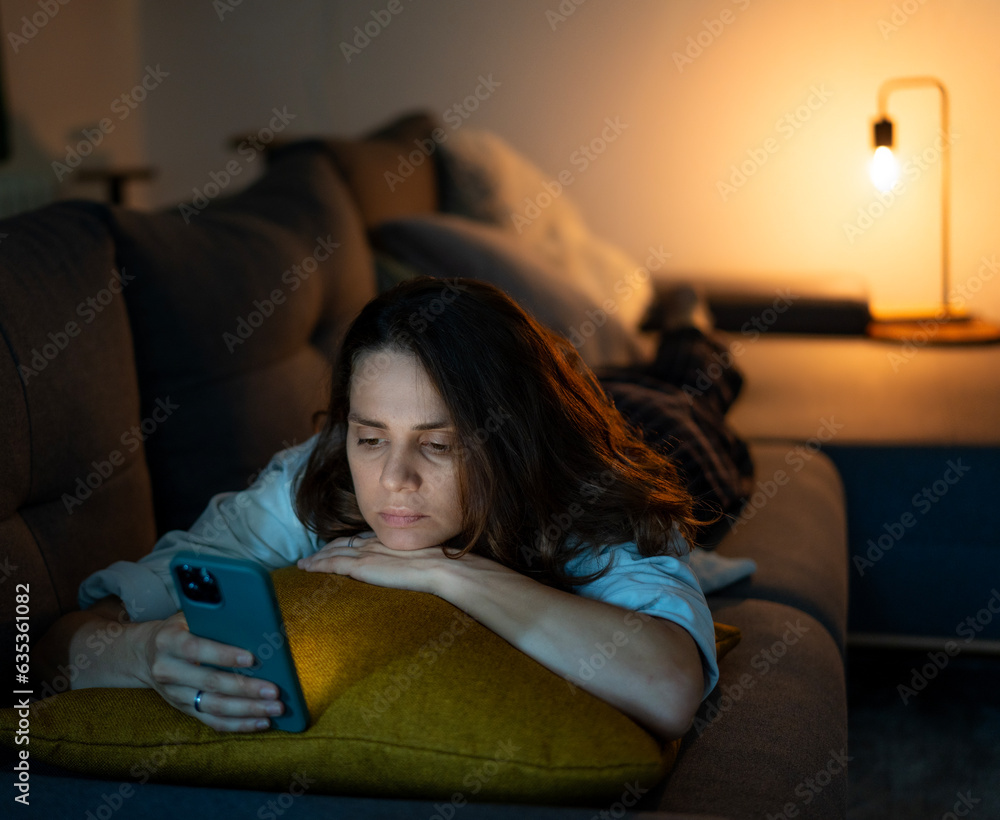Young serious sad woman lying on sofa and looking at smartphone screen at night time at home
