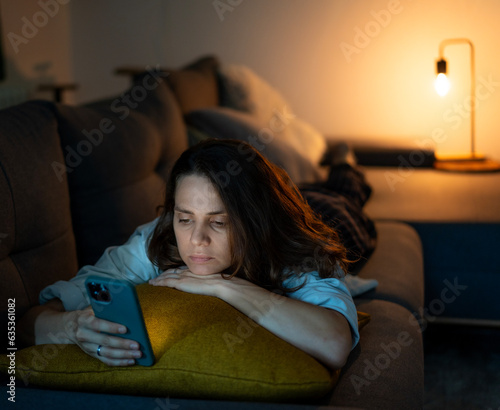 Obraz na płótnie Young serious sad woman lying on sofa and looking at smartphone screen at night time at home w salonie