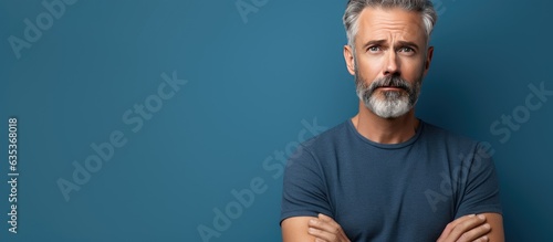 Middle aged man with serious expression standing against blue studio wall looking sad holding back emotions contemplating a problem with empty space