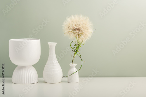Three ceramic vases and a large dandelion on a white table against an olive wall.