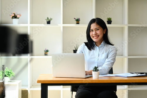Portrait of confident asian female entrepreneur sitting at desk with laptop and smiling to camera.