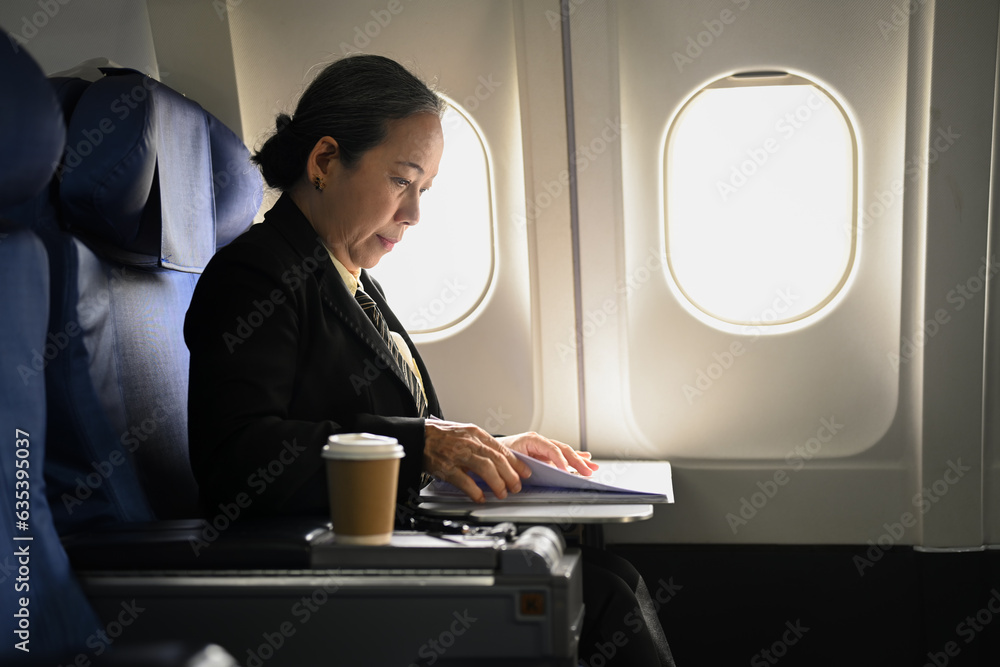Side view of mature businesswoman sitting at her seat in airplane and working with document. Side view of mature businesswoman sitting at her seat in airplane and working with document.
