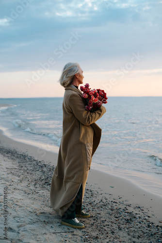 Wall Mural Lonely caucasian middle-aged woman with a bouquet of flowers on seashore in even
