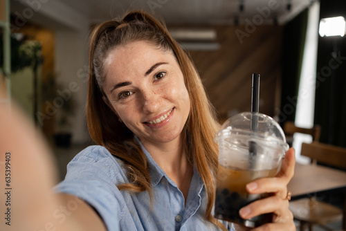 Young freckled woman drinking bubble tea and taking a selfie in a cafe restaurant.