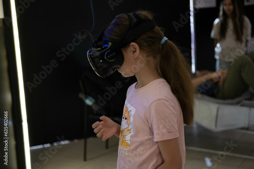Photo of a little girl with long blonde hair in casual clothes, watching videos in modern virtual reality glasses, and playing a game.