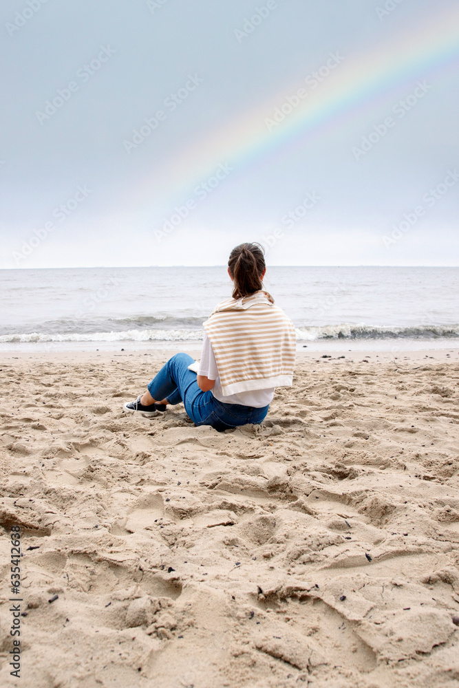 Portrait of a young woman on the beach by the sea, sitting on the seashore. weather after rain, rainbow in the sky. a warm autumn day. rear view