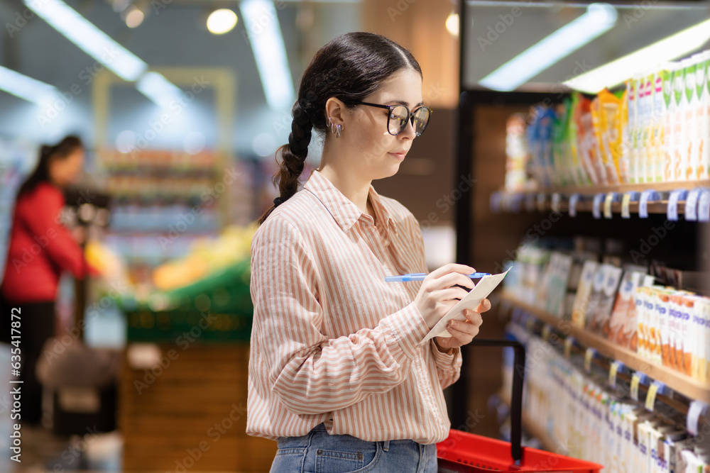 Inspection of grocery store. Portrait of young serious thoughtful caucasian woman wearing eyeglasses writing in document. Concept of shopping in supermarket and consumerism