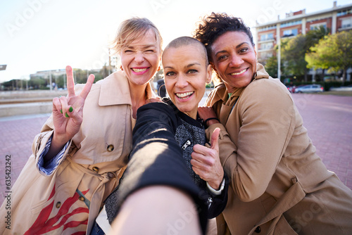 POV selfie mobile of three middle-aged multiracial women looking smiling camera outdoor. Group of cheerful adult friends posing fun for portrait on street on sunny day. Mature friendship relationships photo