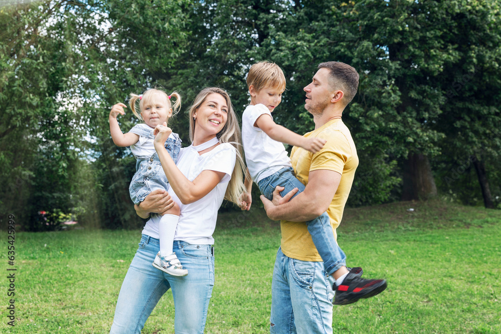 Fototapeta premium Happy family with children in the park on a sunny day. Mom, dad and little daughter and son in jeans and white t-shirts laugh and hug. Love and tenderness.