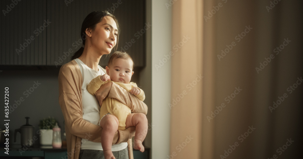 Beautiful Young Asian Mother Holding her Baby in her Arms While Standing Next to a Window at Home. Happy Woman, New to Motherhood, Smiling and Enjoying the Warmth of Sunlight with her Toddler