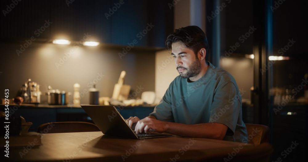 Handsome Authentic Latin Man Sitting at a Table in a Cozy Kitchen and Typing on Laptop Computer at Home at Night. Male Adult Thinking While Browsing the Internet, Checking Videos on Social Networks.