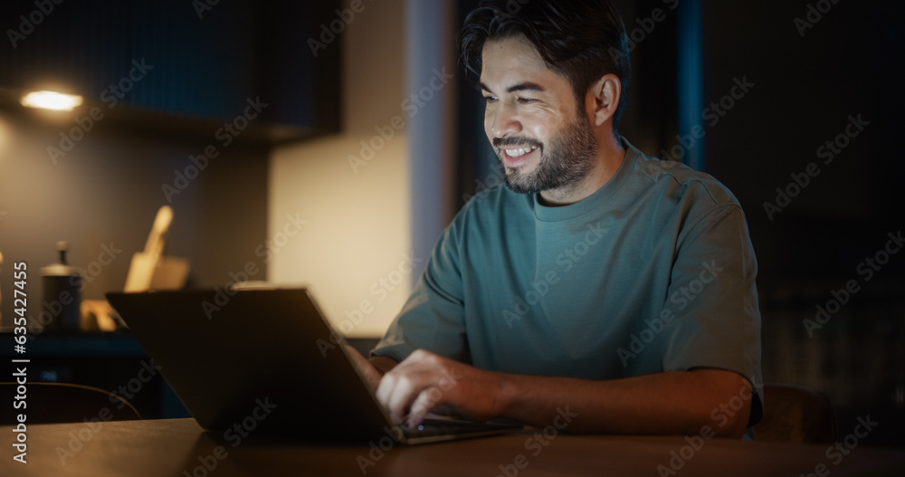 Handsome Authentic Latin Man Sitting at a Table in a Cozy Kitchen and Using Laptop Computer at Home at Night. Male Adult Smiling and Chatting on Social Media, Doing Online Shopping, Checking the News Handsome Authentic Latin Man Sitting at a Table in a Cozy Kitchen and Using Laptop Computer at Home at Night. Male Adult Smiling and Chatting on Social Media, Doing Online Shopping, Checking the News