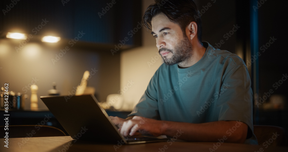 Handsome Authentic Latin Man Sitting at a Table in a Cozy Kitchen and Typing On Laptop Computer at Home at Night. Male Adult Smiling and Chatting on Social Media, Shopping Online, Checking the News. Handsome Authentic Latin Man Sitting at a Table in a Cozy Kitchen and Typing On Laptop Computer at Home at Night. Male Adult Smiling and Chatting on Social Media, Shopping Online, Checking the News.