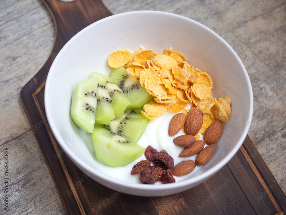 Greek yogurt in a white bowl topped with kiwi, almonds, dried strawberries, and cornflakes, on a wooden cutting board. Top view, flat lay with copy space..
