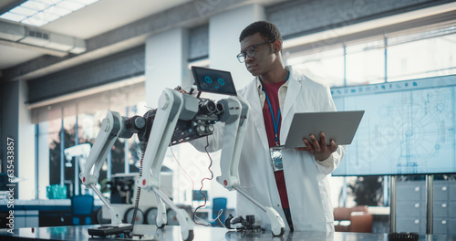Young African American Male Engineer Testing Industrial Programmable Robot Animal in a Factory Development Workshop. Black Researcher in a Lab Coat Developing AI Pet Prototype, Using Laptop