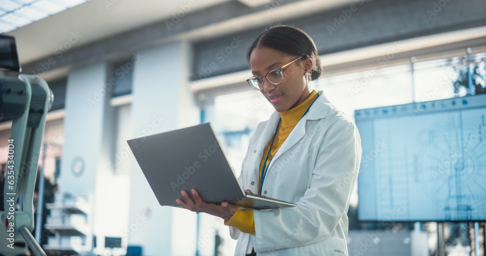 Portrait of Young Black Female Specialist in Lab Coat Using Laptop to Test an AI Robotic Prototype. Professional, Successful Woman Working as an Engineer in Modern High Tech Company Startup Portrait of Young Black Female Specialist in Lab Coat Using Laptop to Test an AI Robotic Prototype. Professional, Successful Woman Working as an Engineer in Modern High Tech Company Startup