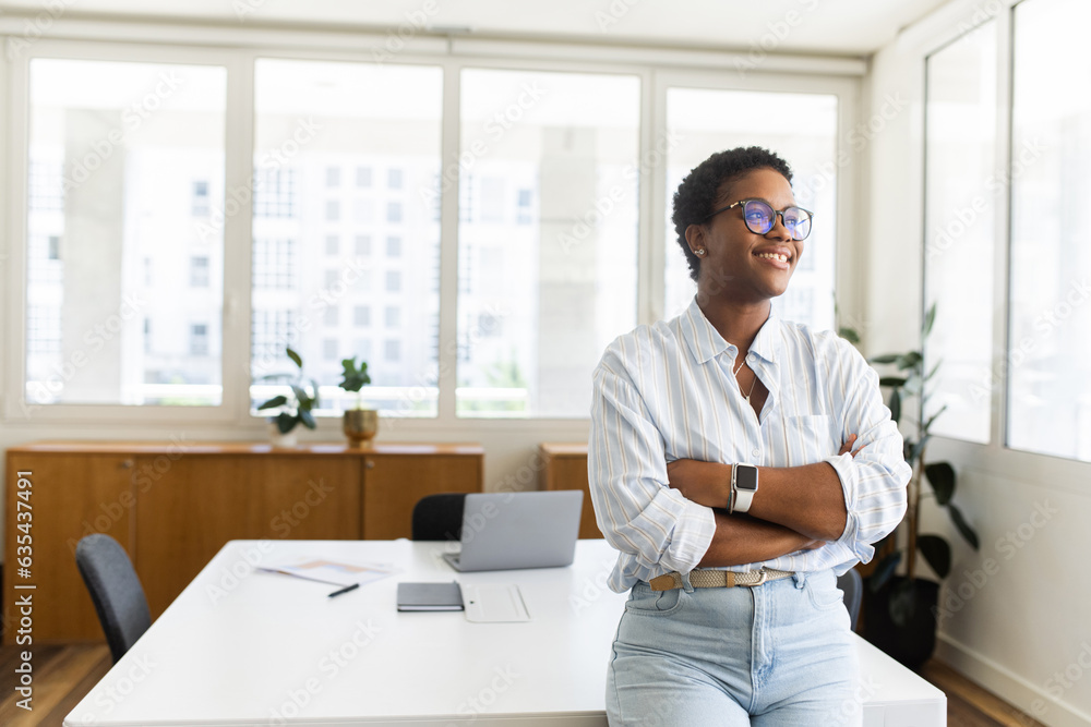 Self-made young African businesswoman, entrepreneur with a short haircut wearing glasses standing alone in a modern office looking away full of creative ideas and plans, determined female freelancer