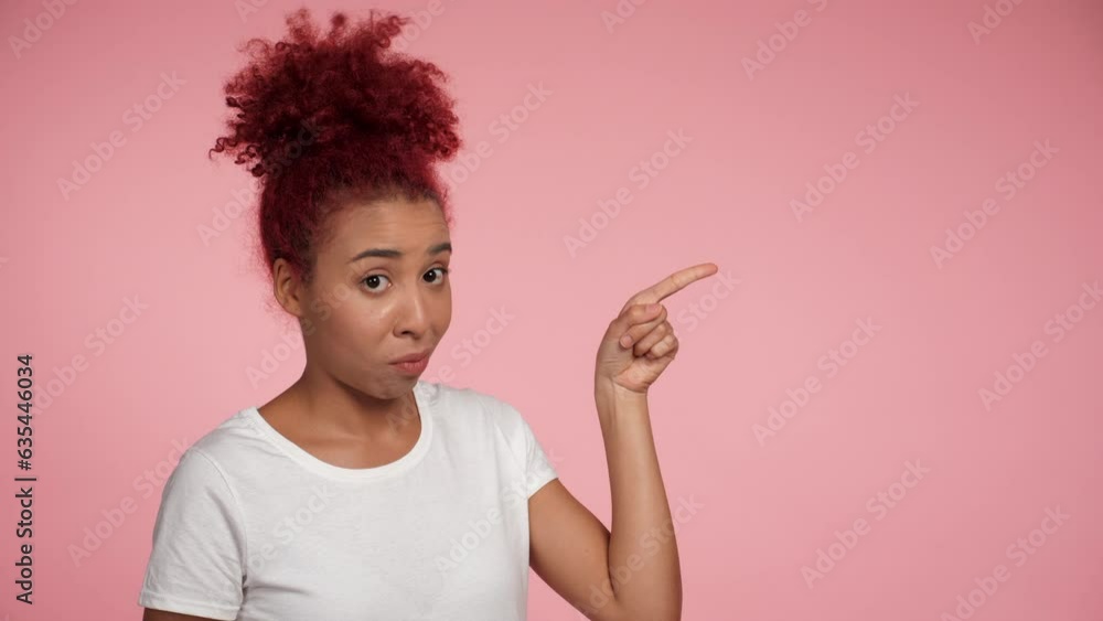Close up cheerful young African American redhead curly woman pointing finger to side workspace promo area mock up and presenting product. Portrait happy female standing on isolated pink background Close up cheerful young African American redhead curly woman pointing finger to side workspace promo area mock up and presenting product. Portrait happy female standing on isolated pink background