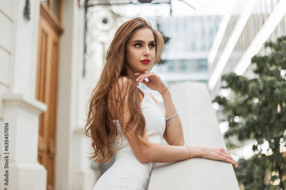 Beautiful young stylish girl with red lips with brunette hair in a fashion elegant white dress stands near a vintage building outdoors Beautiful young stylish girl with red lips with brunette hair in a fashion elegant white dress stands near a vintage building outdoors