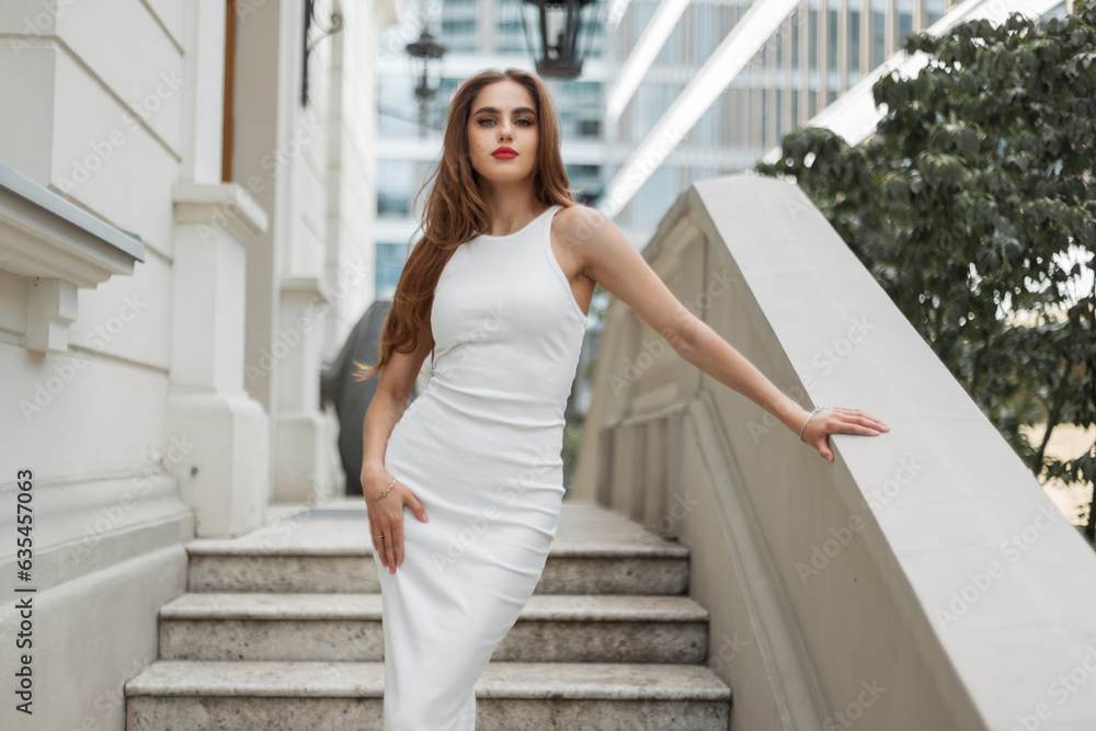 Fashion beautiful woman model with red lips in elegant white dress stands and poses on a vintage steps near a building outdoors Fashion beautiful woman model with red lips in elegant white dress stands and poses on a vintage steps near a building outdoors