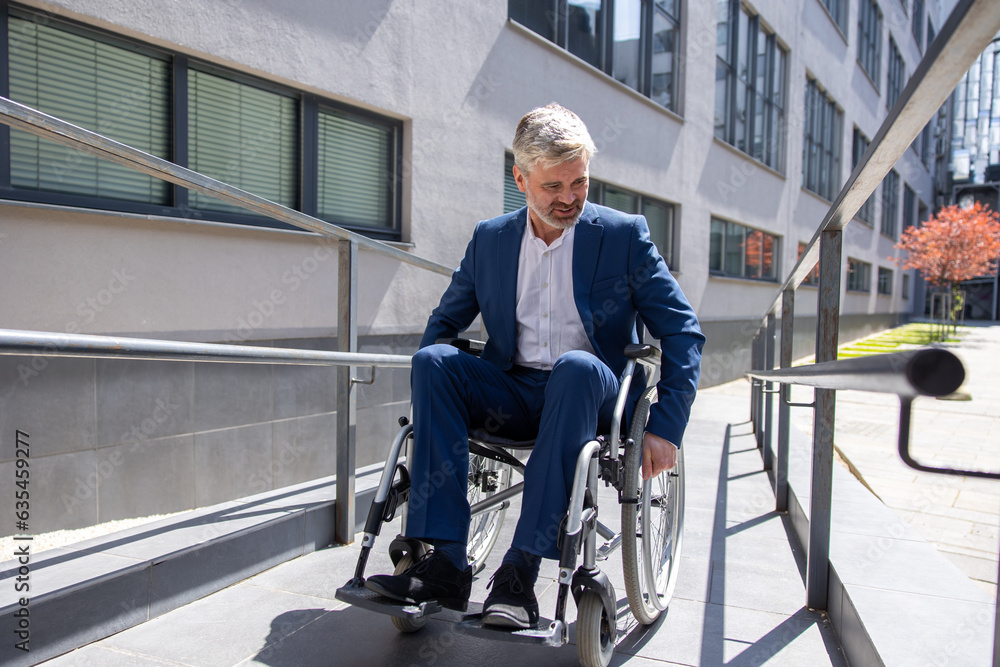 Gray haired businessman in formal wear gets to work in wheelchair rides ramp.