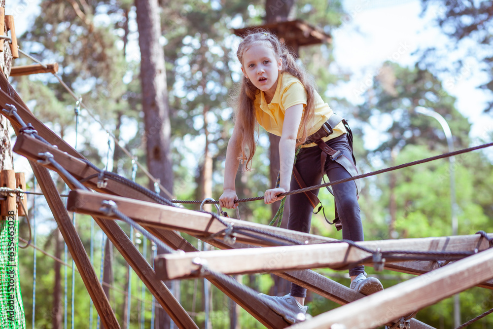 A child in a forest adventure park made of ropes. The girl is climbing the path with a high rope. Children's outdoor climbing entertainment center. Playground for children and sports with a cable car.