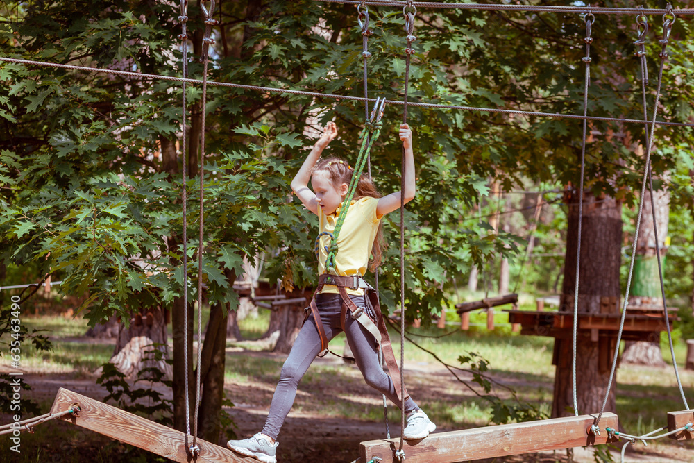 A child in a forest adventure park made of ropes. The girl is climbing the path with a high rope. Children's outdoor climbing entertainment center. Playground for children and sports with a cable car.