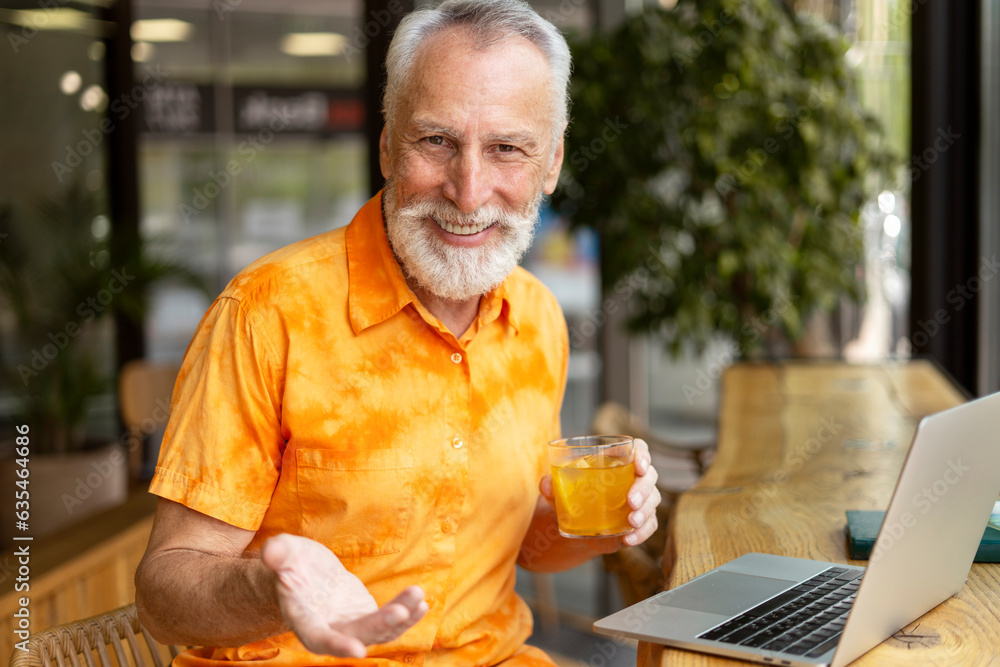 Smiling mature man wearing stylish orange shirt holding lemonade sitting in cafe, using laptop Smiling mature man wearing stylish orange shirt holding lemonade sitting in cafe, using laptop