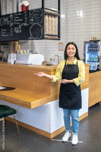 Cute smiling waitress in a black apron standing near the cafe counter photo