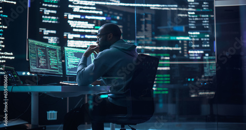 Portrait of a Black Man Working on Computer, Typing Lines of Code that Appear on Big Screens Surrounding him in Monitoring Room. Male Programmer Creating Innovative Software Using AI Data and System