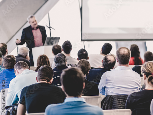 Man giving presentation in lecture hall. Male speeker having talk at public event. Participants listening to lecture. Rear view, focus on people in audience.