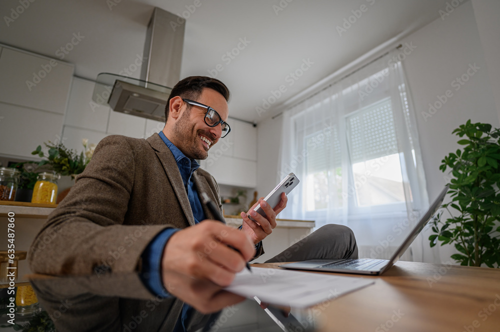 Fototapeta premium Low angle view of smiling businessman in suit using smart phone with laptop on desk at home office