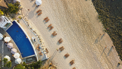 Top view of a beach club with a pool, sunbeds, and umbrellas on a sandy shore by the ocean. White sandy beach, corals and a luxury club on the coast of Bali.