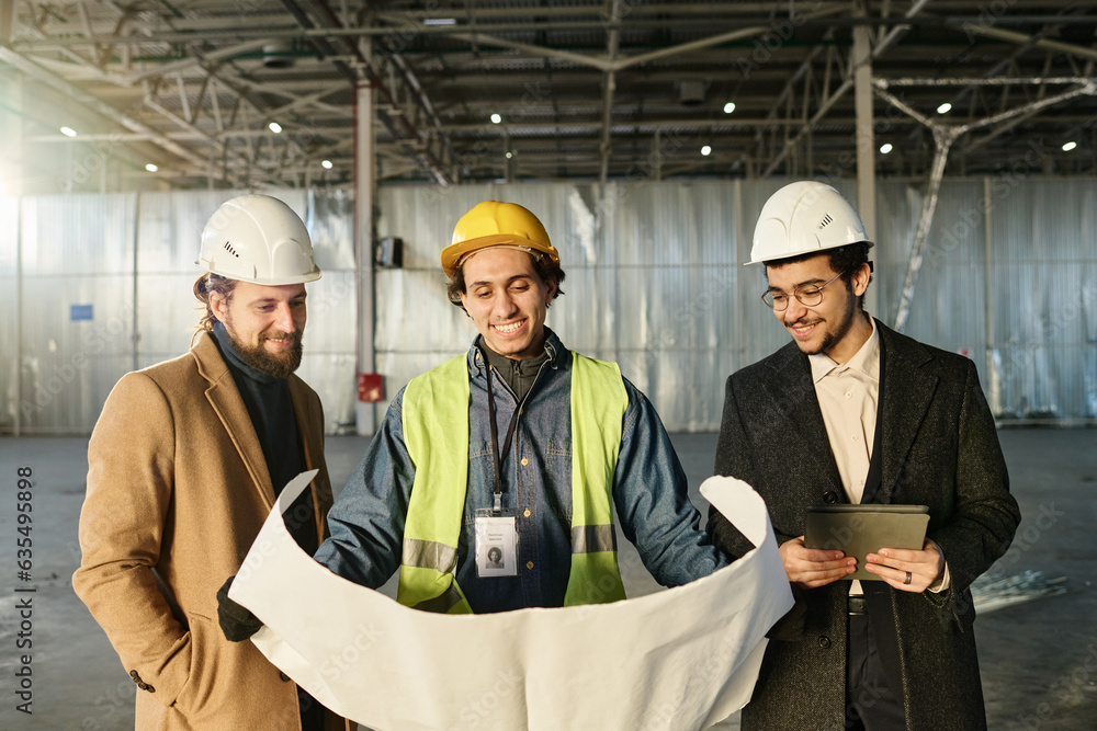 Group of happy young architects in protective helmets looking at sketch of construction or building plan on unrolled blueprint at working meeting