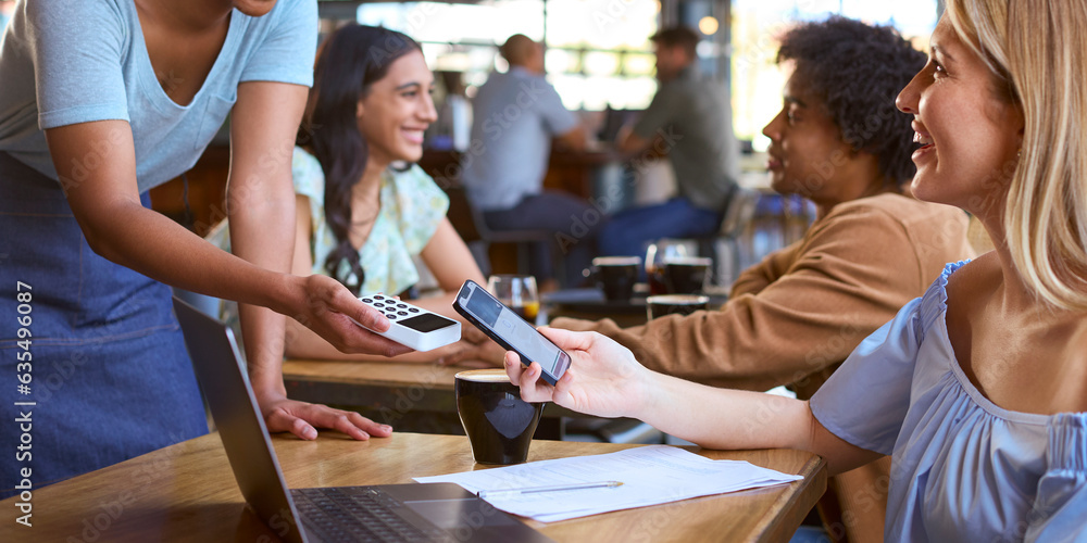 Woman Using Laptop In Coffee Shop Paying Bill With Contactless Mobile Phone Payment Woman Using Laptop In Coffee Shop Paying Bill With Contactless Mobile Phone Payment