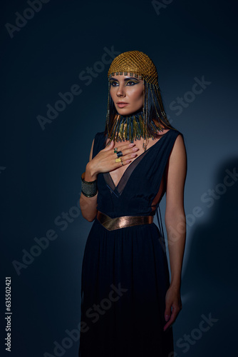 Elegant woman in egyptian headdress and necklace posing while standing on blue background