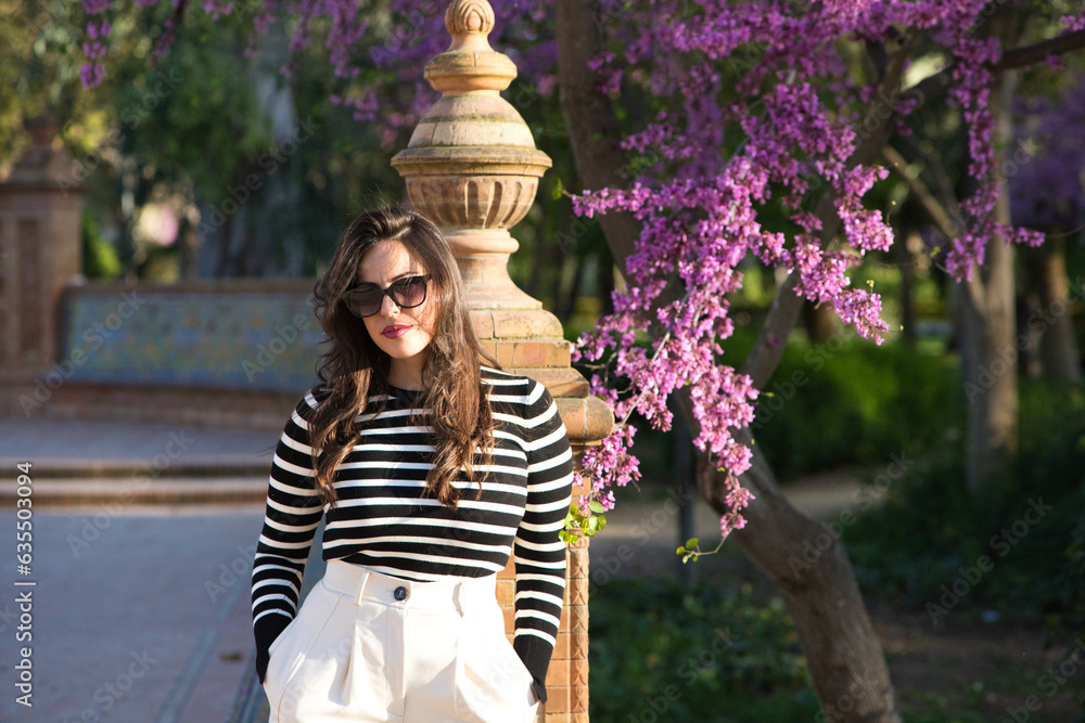 Portrait of a beautiful young woman with long brown hair, wearing a black and white striped shirt, leaning against a column in the park, next to her a tree branch with pink flowers.
