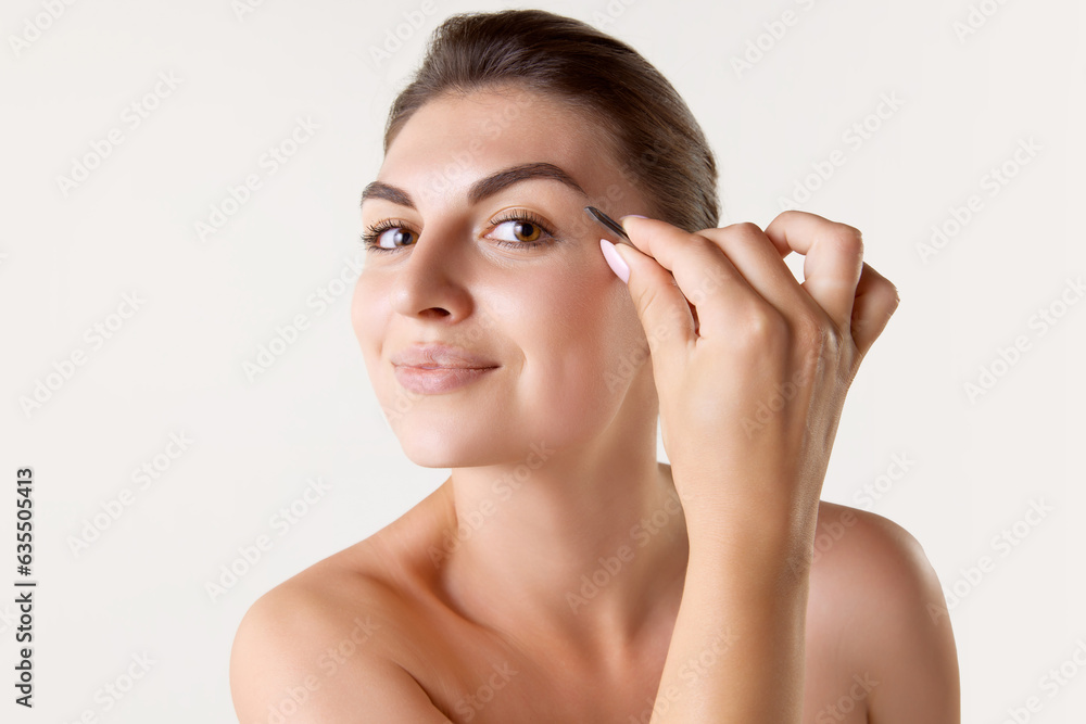 Beautiful young woman plucking eyebrows with tweezers against white studio background. Taking care after appearance. Concept of female beauty, skin care, cosmetology and cosmetics, health, ad