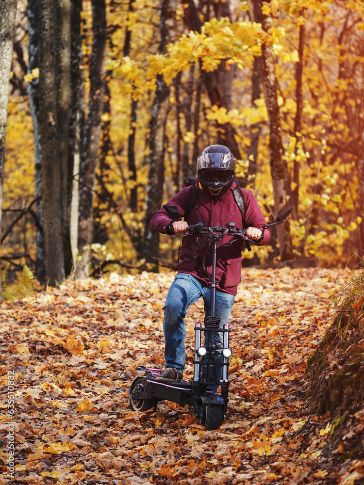 Fototapeta premium A young man rides a scooter in the autumn park. Powerful modern electric transport. Autumn road in maple leaves