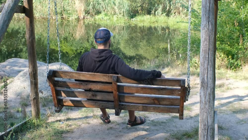 a bearded man in a cap resting on the wooden swing near a pond a bearded man in a cap resting on the wooden swing near a pond