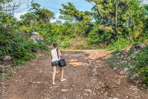 A young female tourist with a camera bag climbs a forest road in the mountains near Budva, Montenegro. Hiking among summer nature