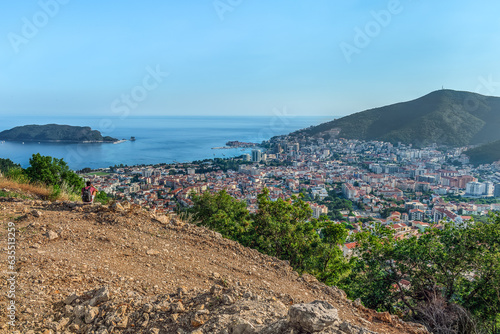 View from the top of the mountain to the city of Budva down in the valley on the Adriatic Sea, Montenegro. A young man with a backpack, squatting, looks at the seascape with Sveti Nikola Island