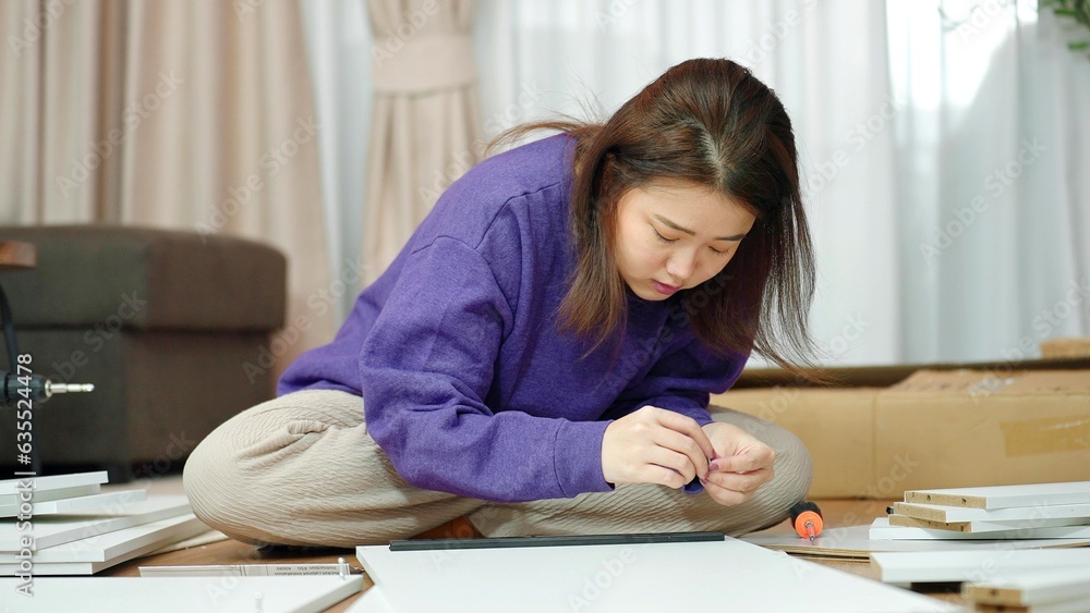 Young Asian woman sitting on floor in living room while assembling furniture at home. Asian woman assembling furniture by herself. Assembling furniture concept Young Asian woman sitting on floor in living room while assembling furniture at home. Asian woman assembling furniture by herself. Assembling furniture concept