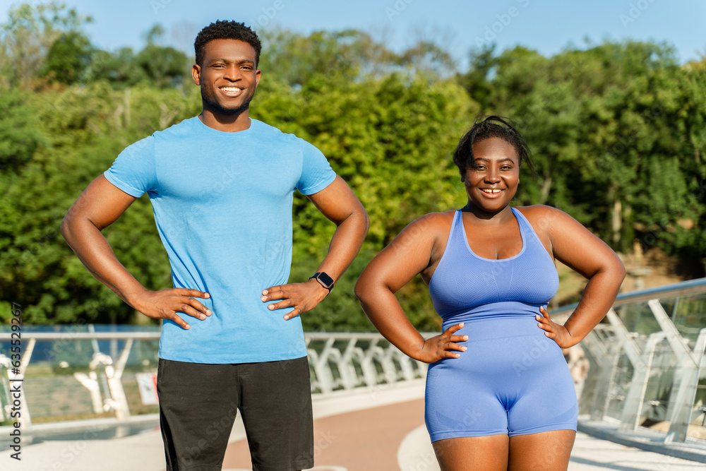 Portrait of smiling African American man and woman doing exercises in park. Happy plus size model training with personal trainer on the street. Sport, motivation, healthy lifestyle concept