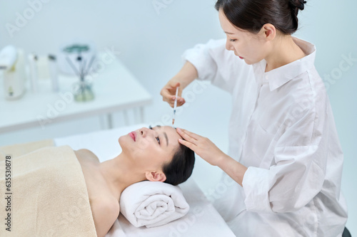 아시아 한국의 젊은 여성이 피부과 병원 또는 성형외과의 침대에 누워 베이지색 대형 타올을 덮고 눈을 감고 있고 하얀색 셔츠를 입은 전문의사가 주사기를 들고 충전제 또는보톡스로 노화방지 또는 얼굴 주름을 관리하고 있다. 