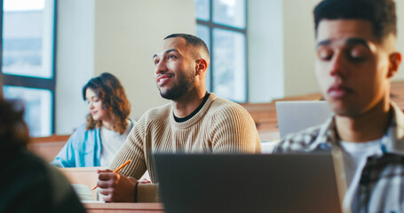 Handsome young African American male student sitting among others, listening to professor and noting information at lection. High school concept. Guy studying at University and writing down.