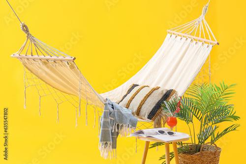 Cozy hammock with plant, beach accessories and cocktail on stool against yellow background. Summer vacation concept