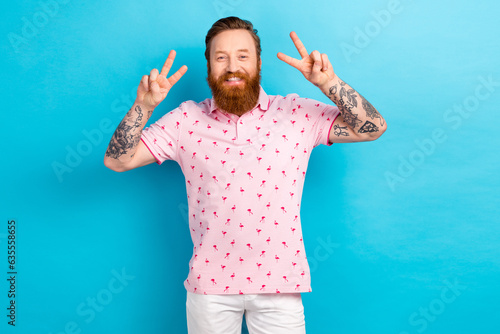 Portrait of good mood satisfied man with cool tattoo wear flamingo t-shirt showing v-sign greetings isolated on blue color background