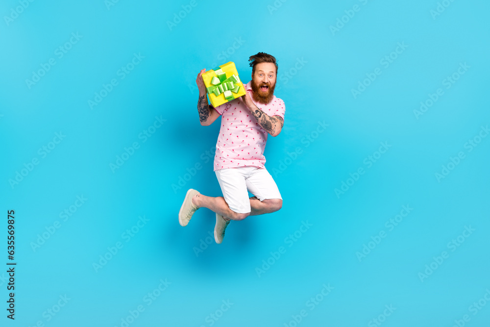 Full length photo of overjoyed young man jumping hands hold desirable giftbox isolated on blue color background Full length photo of overjoyed young man jumping hands hold desirable giftbox isolated on blue color background