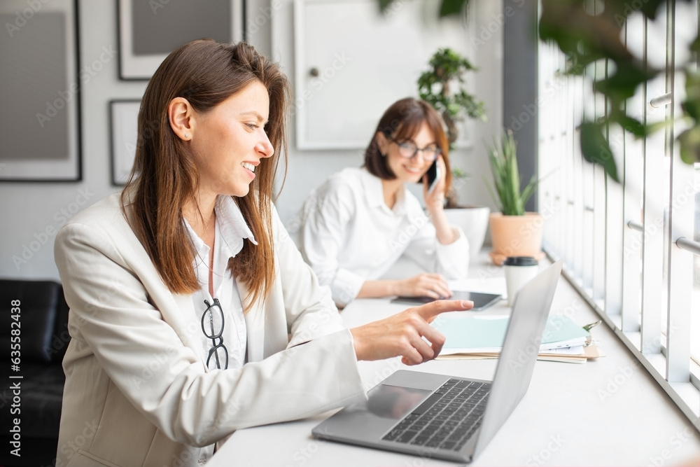 Female company employees working in office lady pointing at laptop screen while colleague talking on phone on background Female company employees working in office lady pointing at laptop screen while colleague talking on phone on background
